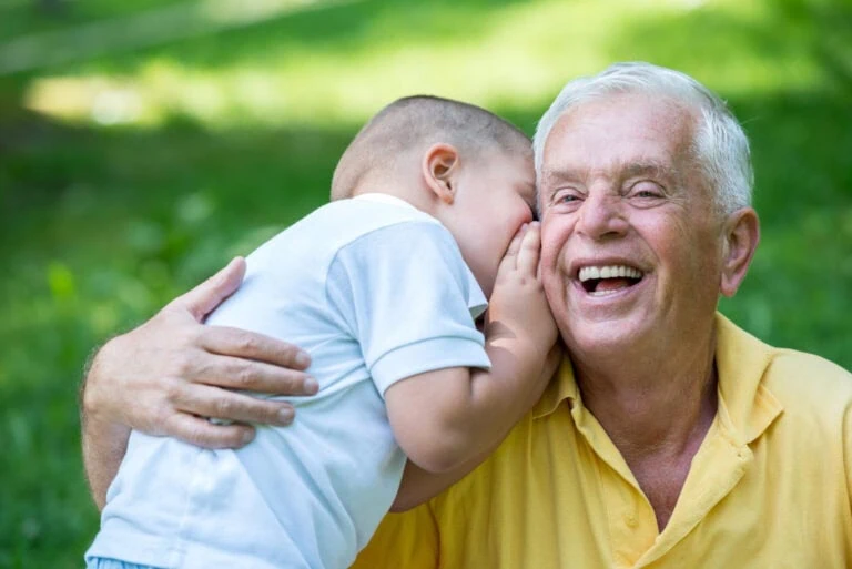 Elderly man smiling as grandson whispers a secret in his ear in a park setting.